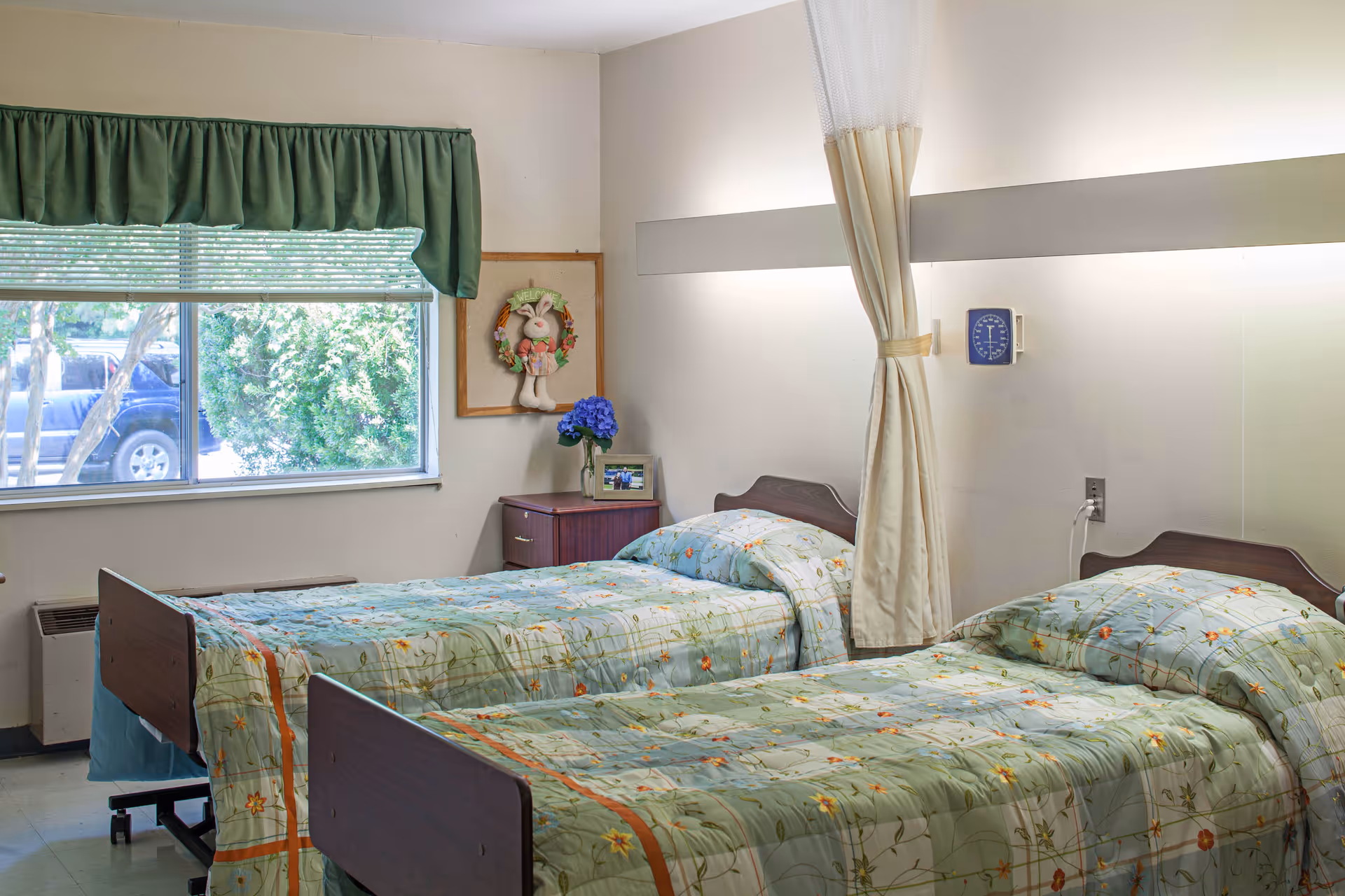 A shared bedroom in a senior living facility with two single beds covered in floral patterned bedspreads. There is a window with green curtains and a view of trees and a parked car outside. A small wooden nightstand between the beds holds a vase with blue flowers and a framed photo. A wall-mounted clock and a privacy curtain are also visible.