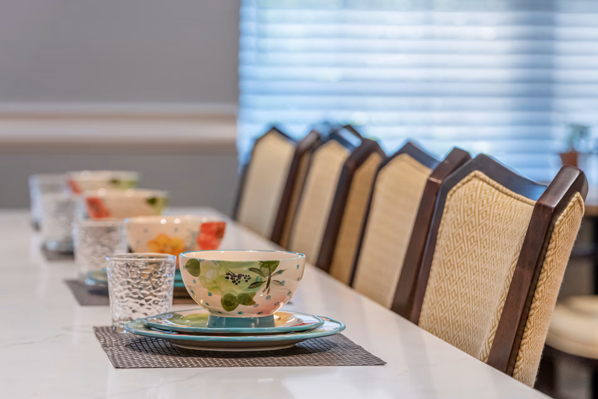 Place settings with floral bowls, plates, and glasses arranged along a countertop with upholstered dining chairs in the background.