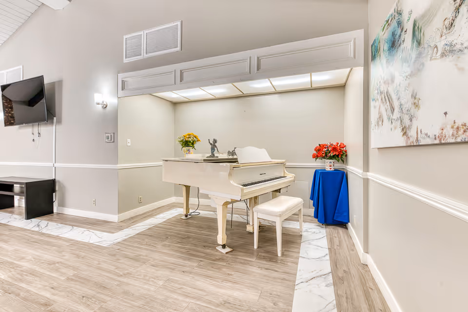 Bright common room featuring a white baby grand piano with a bench, wall-mounted TV, flowers, and artwork.