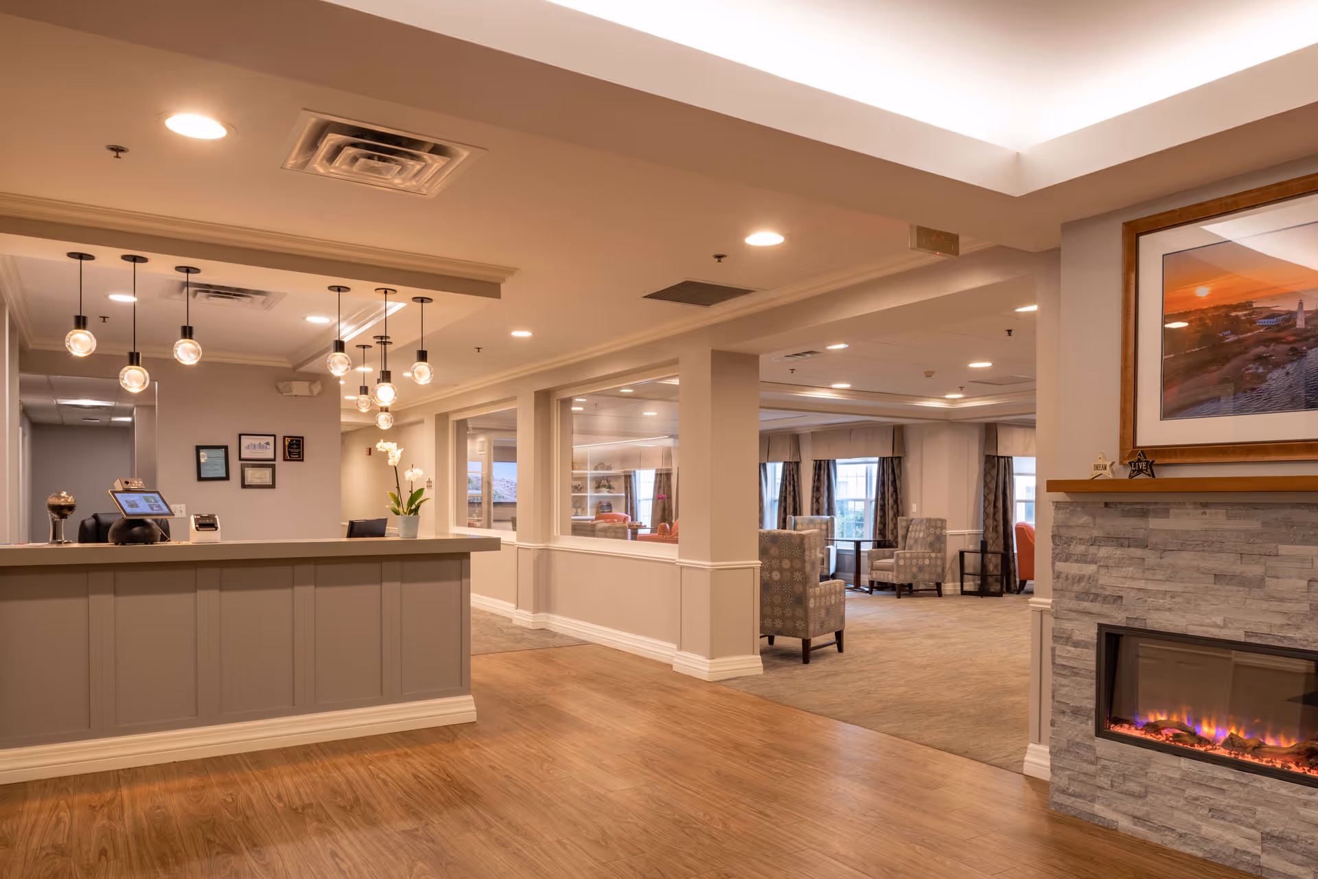 Interior view of a senior living facility reception area with a front desk, hanging pendant lights, and a seating area with armchairs and large windows in the background. There is a modern electric fireplace with a framed picture above it on the right side.