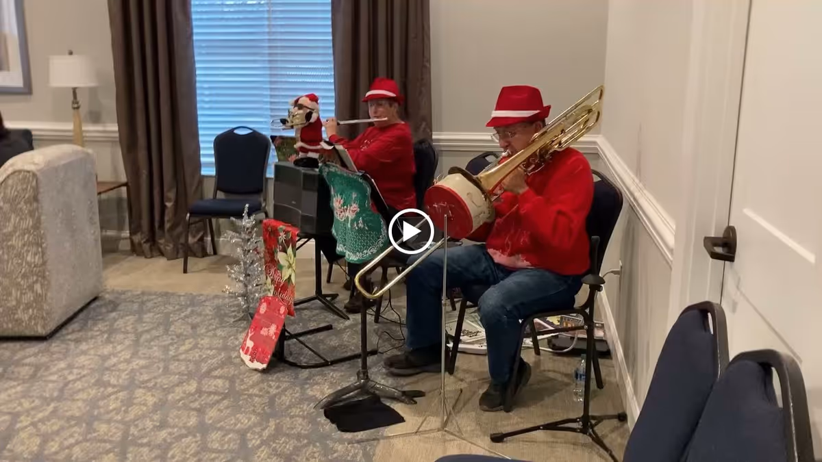 Two elderly individuals wearing red sweaters and red hats are seated in a room playing musical instruments, one playing a trombone and the other a flute. The room has carpeted flooring, a window with closed blinds and brown curtains, a lamp, and several chairs. There are festive holiday decorations including a small silver Christmas tree and a Santa Claus figure on a music stand.