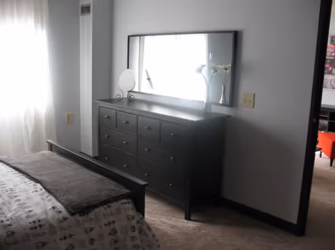 Bedroom with a dark wood dresser and large mirror, a bed in the foreground, and light from a curtained window.