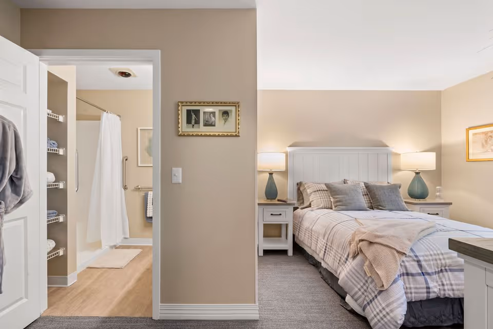 A cozy bedroom with a neatly made bed featuring plaid bedding and multiple pillows. Two bedside tables with matching lamps flank the bed. The walls are painted beige and decorated with framed pictures. To the left, an open door reveals a bathroom with a shower curtain, grab bars, and shelves stocked with towels.