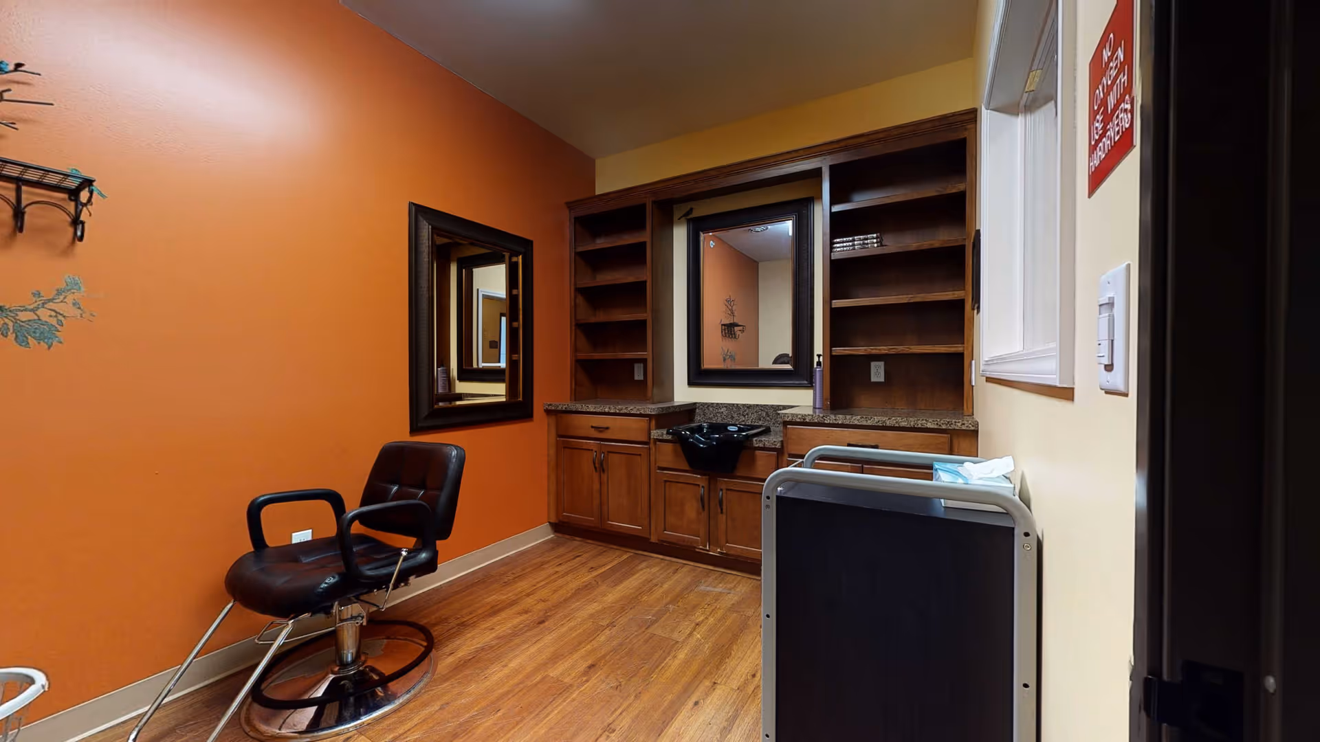 Interior view of a salon area in a retirement living facility with an orange accent wall, a black salon chair, a large mirror, wooden cabinets with a black sink, and open shelving.