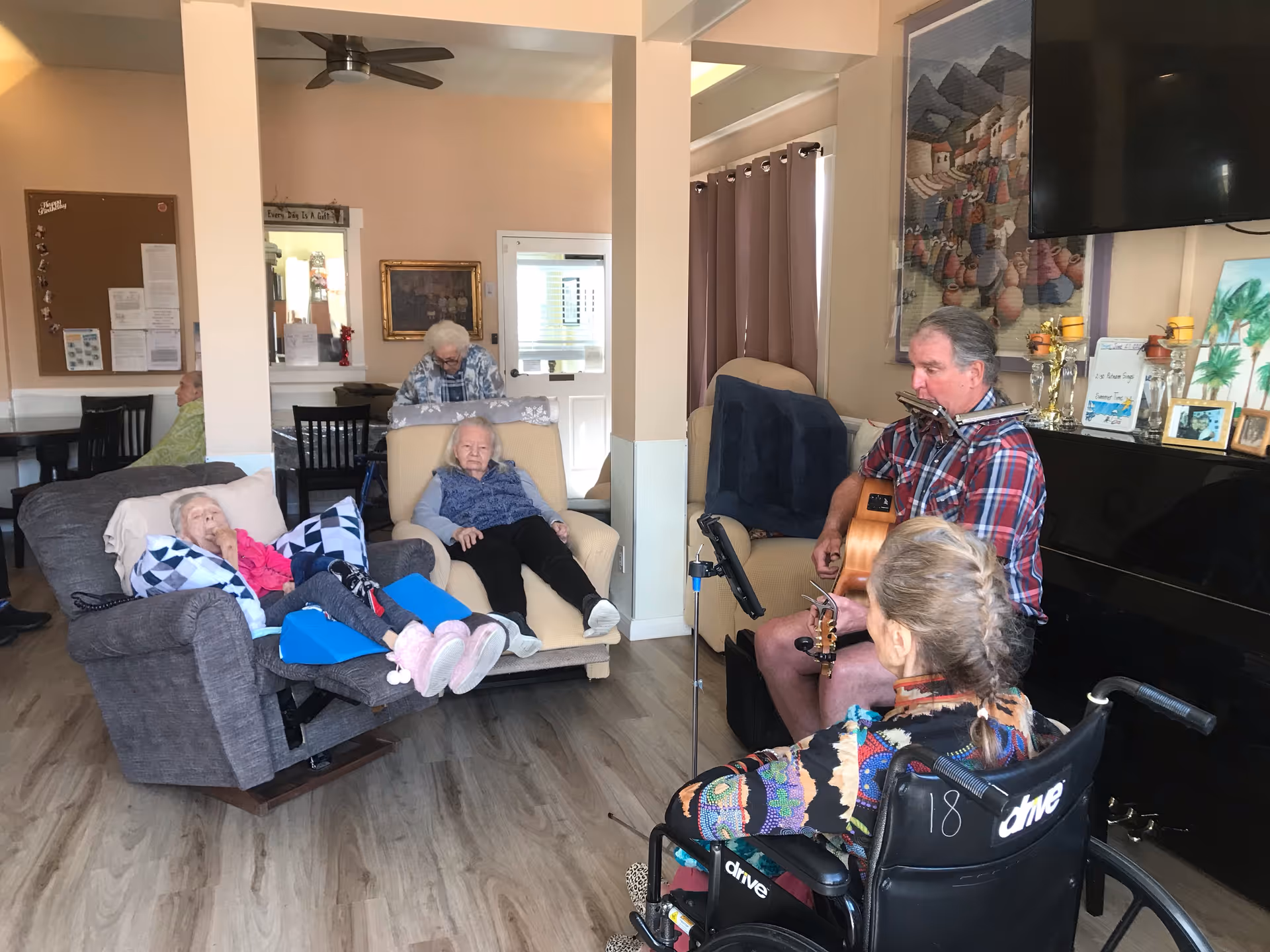 Residents in a senior living common room listening to a man playing guitar and harmonica.