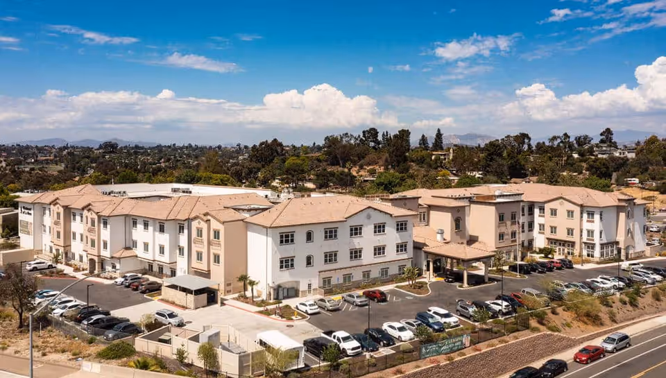 Aerial view of a large, multi-story senior living facility with beige and white exterior walls and a tan roof. The building is surrounded by parking lots filled with cars, landscaped areas, and a backdrop of trees and distant hills under a partly cloudy blue sky.