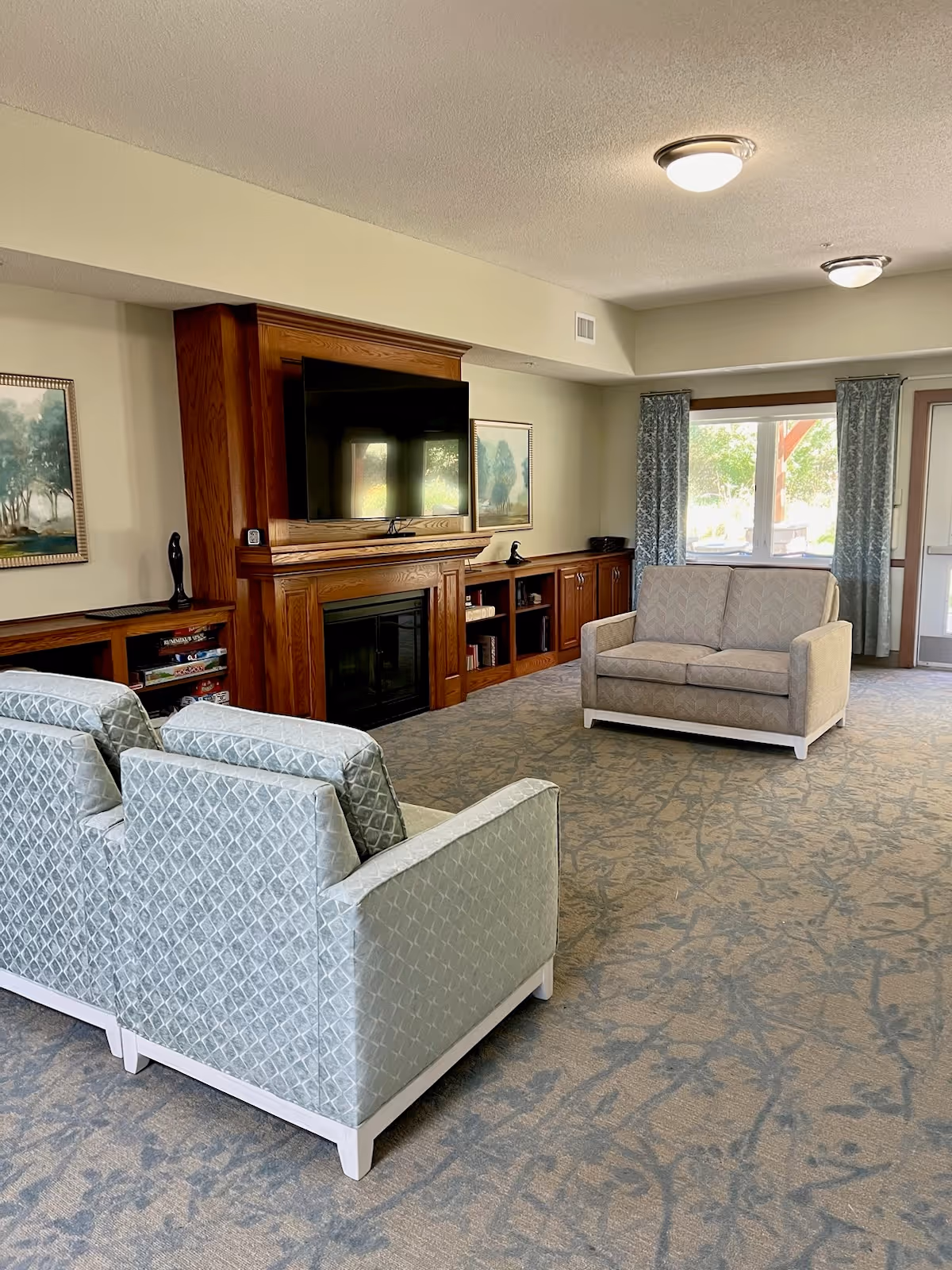 Spacious communal living room with upholstered chairs and a loveseat facing a wall-mounted TV above a wooden fireplace, with windows and curtains in the background.