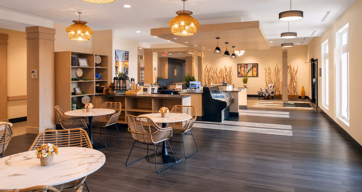 Bright communal dining/lounge area with round marble-top tables and wicker chairs facing a service counter and reception desk in a modern senior living facility.