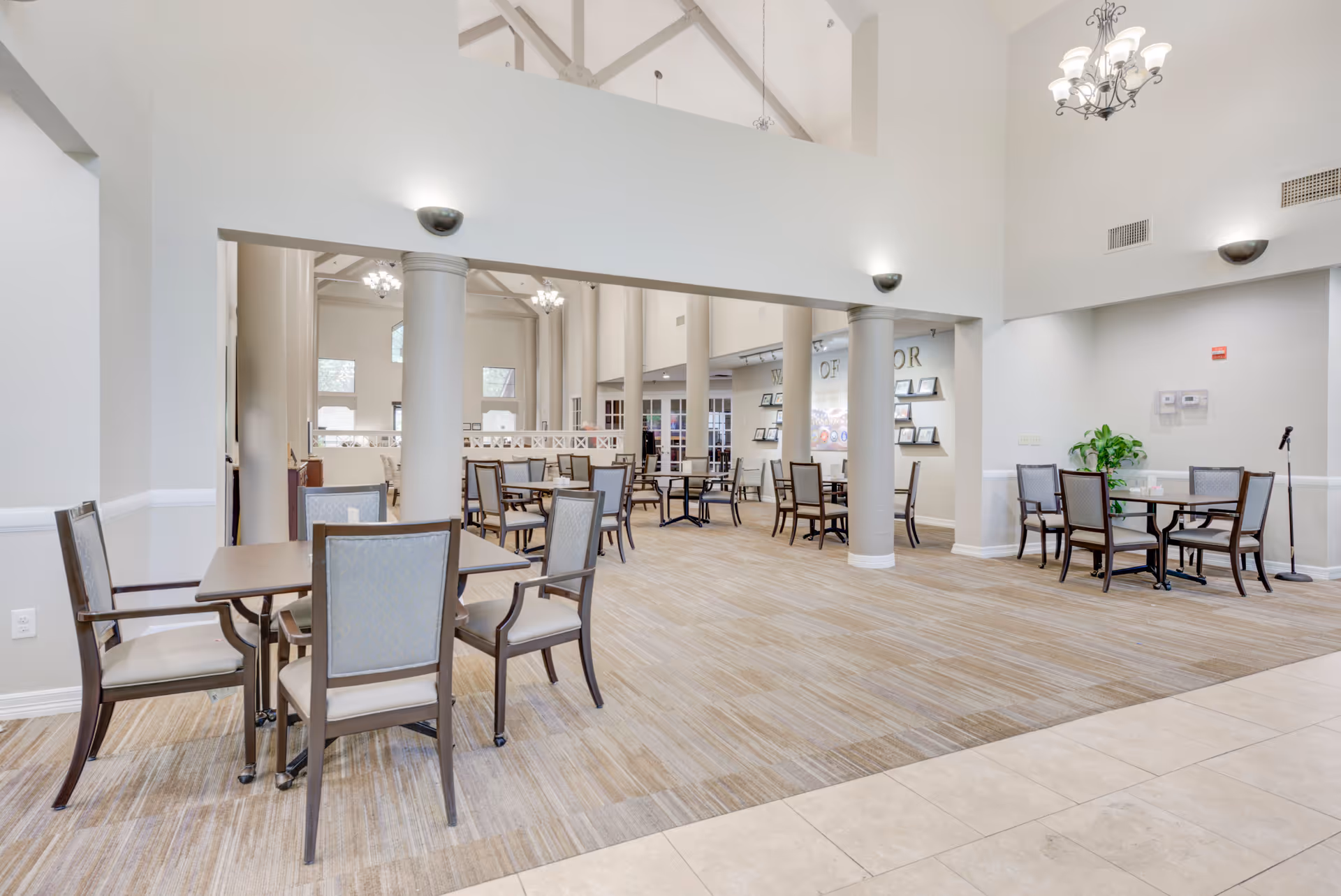 A spacious and well-lit dining area in The Continental Retirement Community featuring multiple tables with chairs arranged neatly. The room has high ceilings with chandeliers, large columns, and a carpeted floor with a tiled section in the foreground. There are framed pictures on the walls and a plant in the corner.