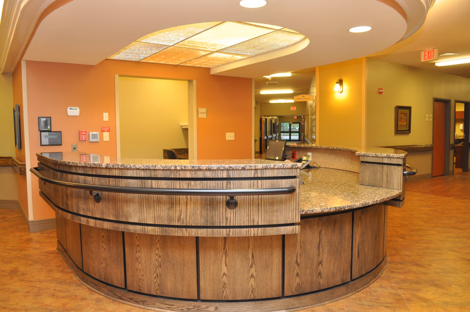 Curved wooden reception desk with granite countertop in a well-lit hallway of a senior living facility. The walls are painted in warm tones of orange and yellow, with a ceiling light fixture above the desk. The hallway extends into the background with doors and framed artwork on the walls.