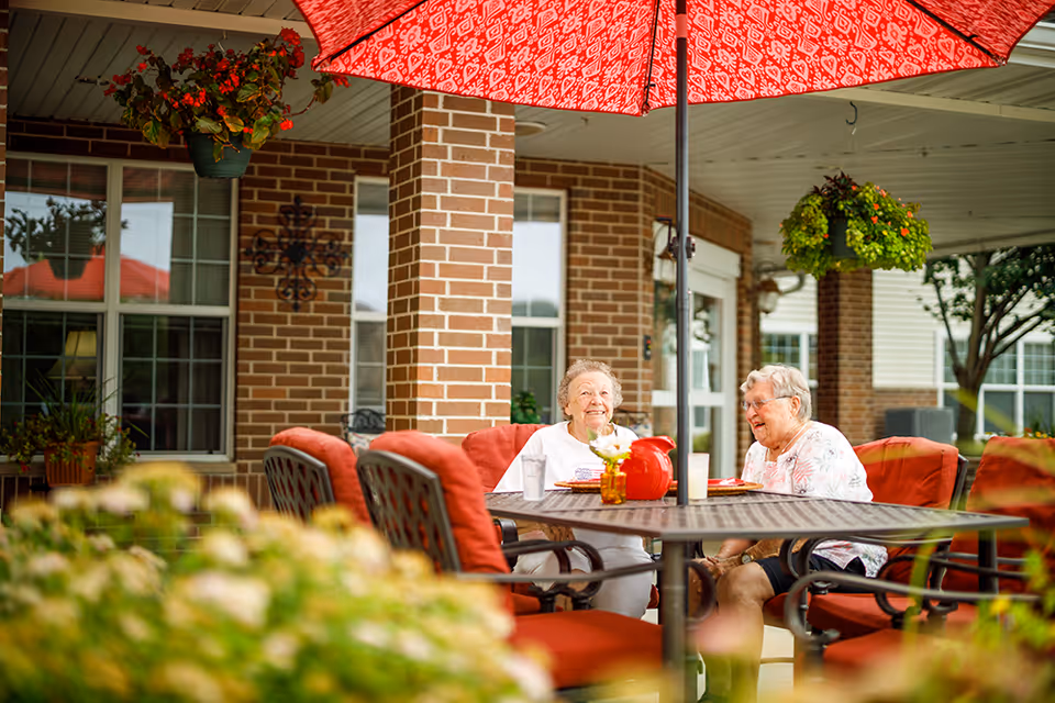 Two elderly women sitting and smiling at a table with red cushioned chairs under a large red umbrella on a covered patio with brick pillars and hanging flower baskets.