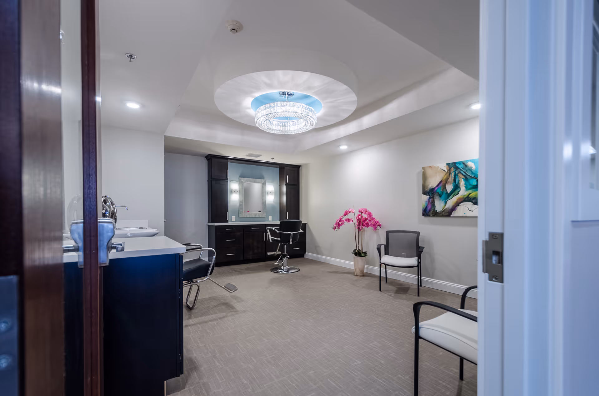 Interior view of a well-lit salon area in a senior living facility with a modern chandelier on the ceiling, two salon chairs, a large mirror with cabinetry, two guest chairs, a potted orchid plant, and abstract wall art.
