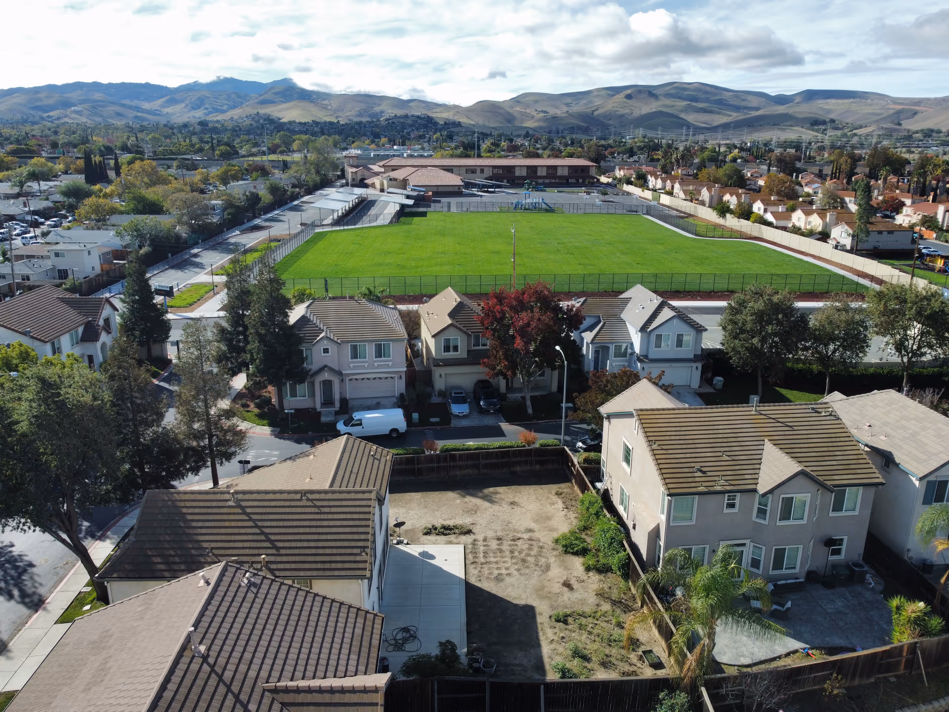 Aerial view of a residential neighborhood with several houses, a large green sports field surrounded by a fence, and hills in the background under a partly cloudy sky.
