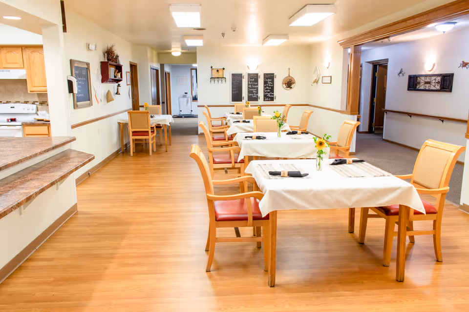 Bright dining room in a senior living facility with several tables and chairs set with tablecloths, place settings, and small flower vases, with a serving counter/kitchen area to the left.
