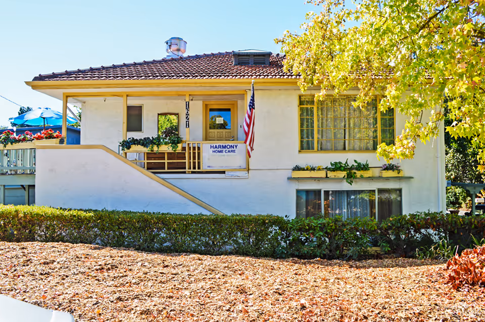 Exterior view of a single-story building with a tiled roof and a small porch. The porch has a railing with plants and an American flag next to the entrance door. A sign on the porch reads 'Harmony Home Care'. There are windows with curtains and flower boxes, and a tree with green and yellow leaves partially shading the building. The ground in front is covered with dry leaves and bushes.