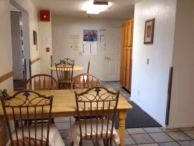 Interior view of a dining area in a senior living facility with a wooden table and four chairs in the foreground, another small round table with two chairs in the background, white walls, tile flooring, a door, and wooden cabinets along the right wall.