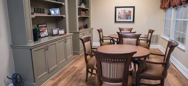 Small dining room with round wooden tables and upholstered chairs, built-in shelving and cabinets, framed artwork, and windows.