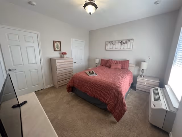 A bedroom with a bed covered in a red quilt and matching pillows. There is a wooden dresser with a small framed picture and a vase with flowers on top. Two closed white doors are visible on the left wall. A nightstand with a lamp is on each side of the bed. A window with blinds is on the right wall, next to an air conditioning unit. A TV is partially visible in the foreground on the left side.