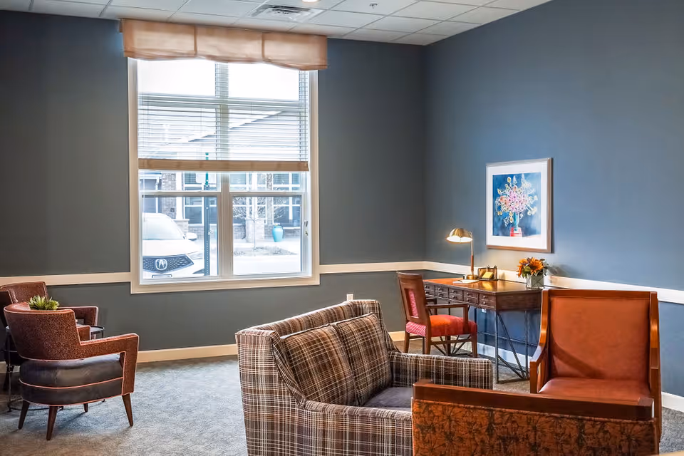 Cozy lounge with armchairs, a plaid loveseat, a writing desk with lamp and a large window.