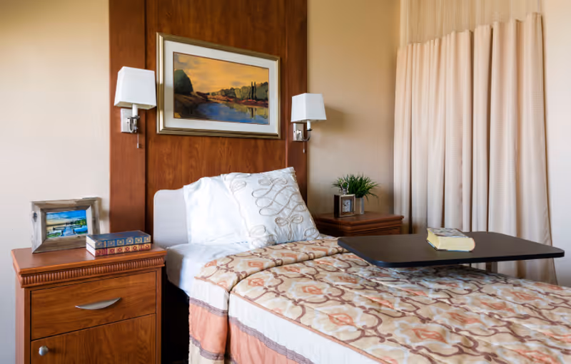 A neatly made single bed with a patterned bedspread and white pillows in a senior living facility room. The bed has a wooden headboard with two wall-mounted lamps and a framed landscape painting above it. There are two wooden nightstands on either side of the bed, one with a small framed picture and books, and the other with a small plant and a clock. A beige curtain is partially visible on the right side, and a black overbed table with a book on it is positioned over the bed.