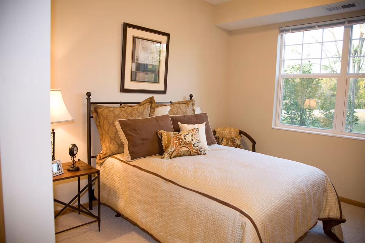A cozy bedroom with a neatly made bed featuring a cream-colored quilt and multiple decorative pillows in brown and patterned designs. There is a wooden side table with a lamp, a small clock, and a framed photo. A framed abstract artwork hangs on the wall above the bed. A window on the right side lets in natural light and shows greenery outside. A wicker chair with a cushion is placed in the corner near the window.