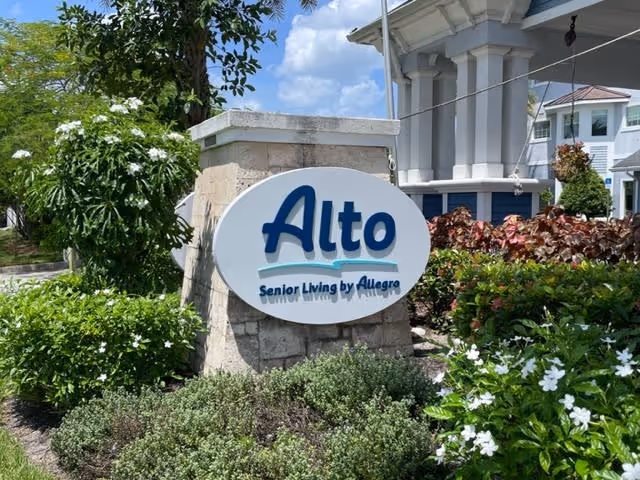 Outdoor view of a stone sign for Alto Senior Living by Allegro surrounded by green bushes and white flowers, with part of a building and blue sky with clouds in the background.