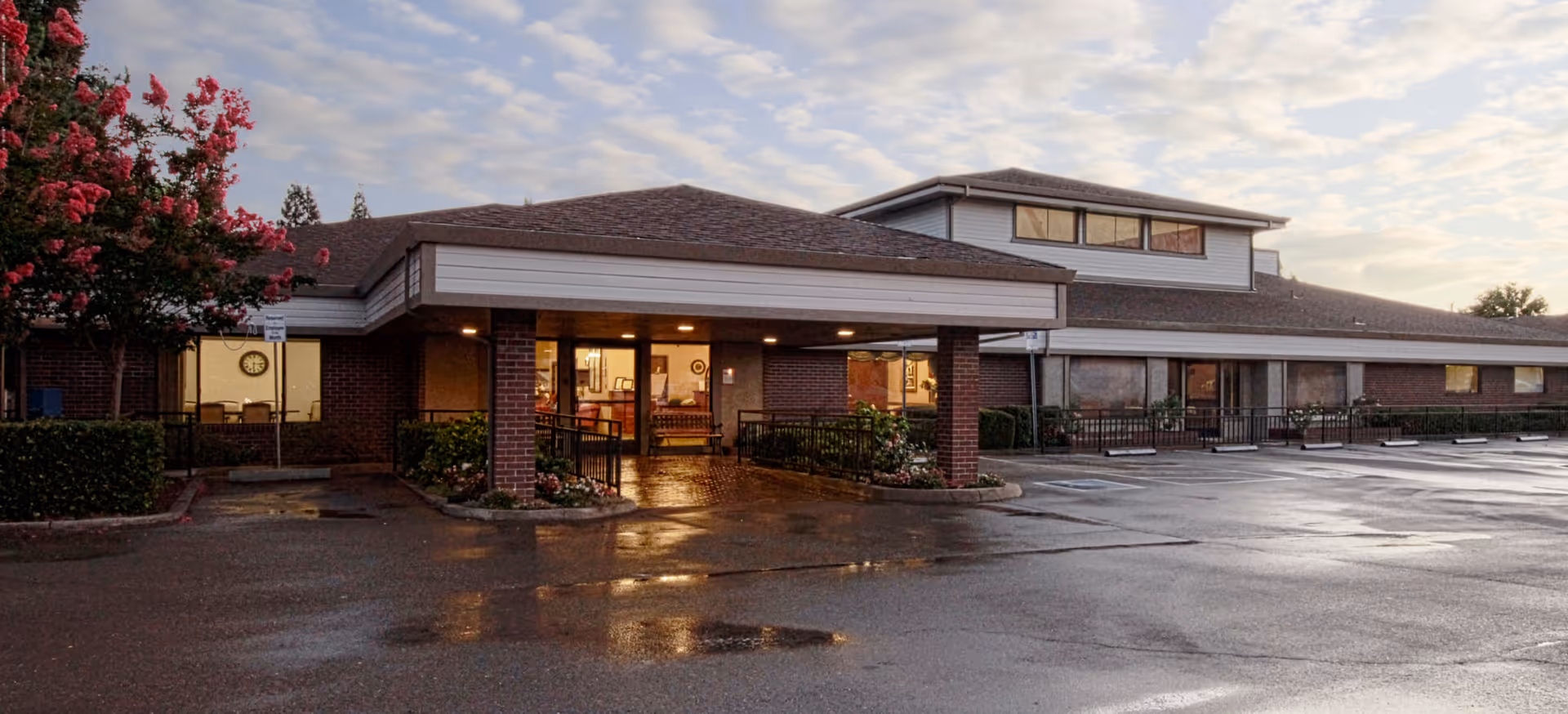 Exterior view of Pine Creek Care Center at dusk, showing a single-story brick building with a covered entrance, large windows, and a wet parking lot reflecting the evening light. There are flowering trees and shrubs near the entrance.
