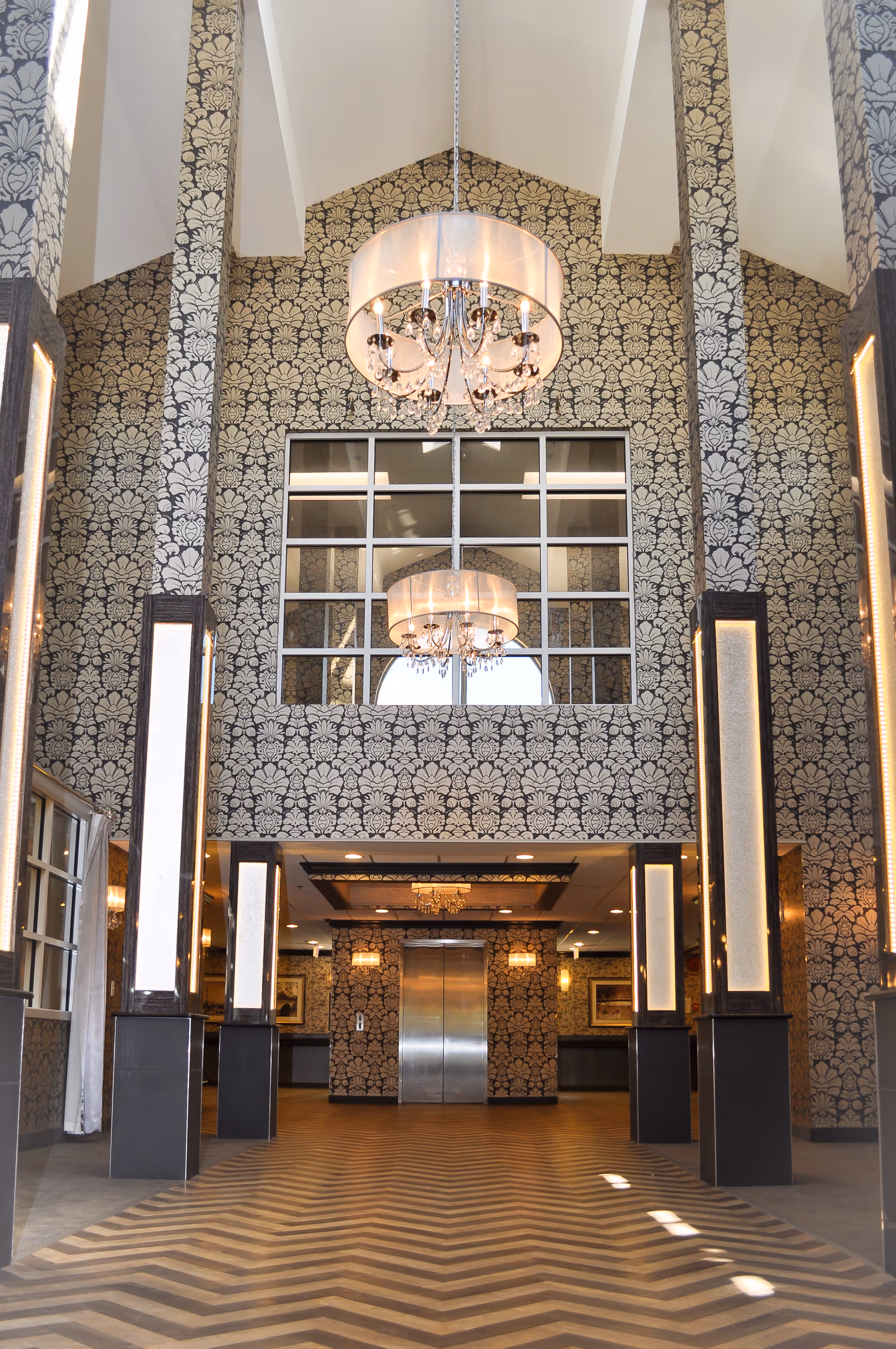 Interior view of a spacious lobby area with patterned wallpaper, a large chandelier hanging from a high ceiling, and an elevator at the far end. The floor has a chevron pattern, and tall illuminated columns line the sides of the space.
