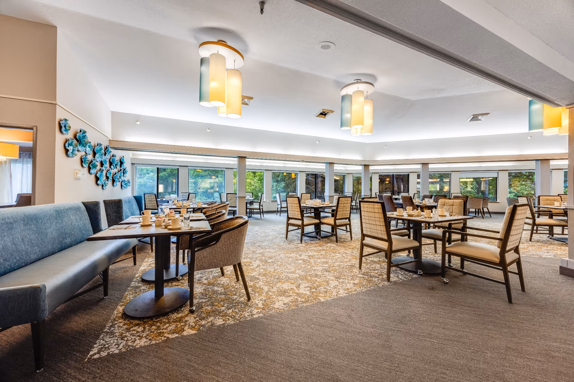 A spacious dining room with multiple tables and chairs arranged neatly. The room features large windows allowing natural light to brighten the space, modern pendant lights hanging from the ceiling, and a blue cushioned bench along one wall with decorative blue wall art above it. Tables are set with cups, glasses, and utensils.