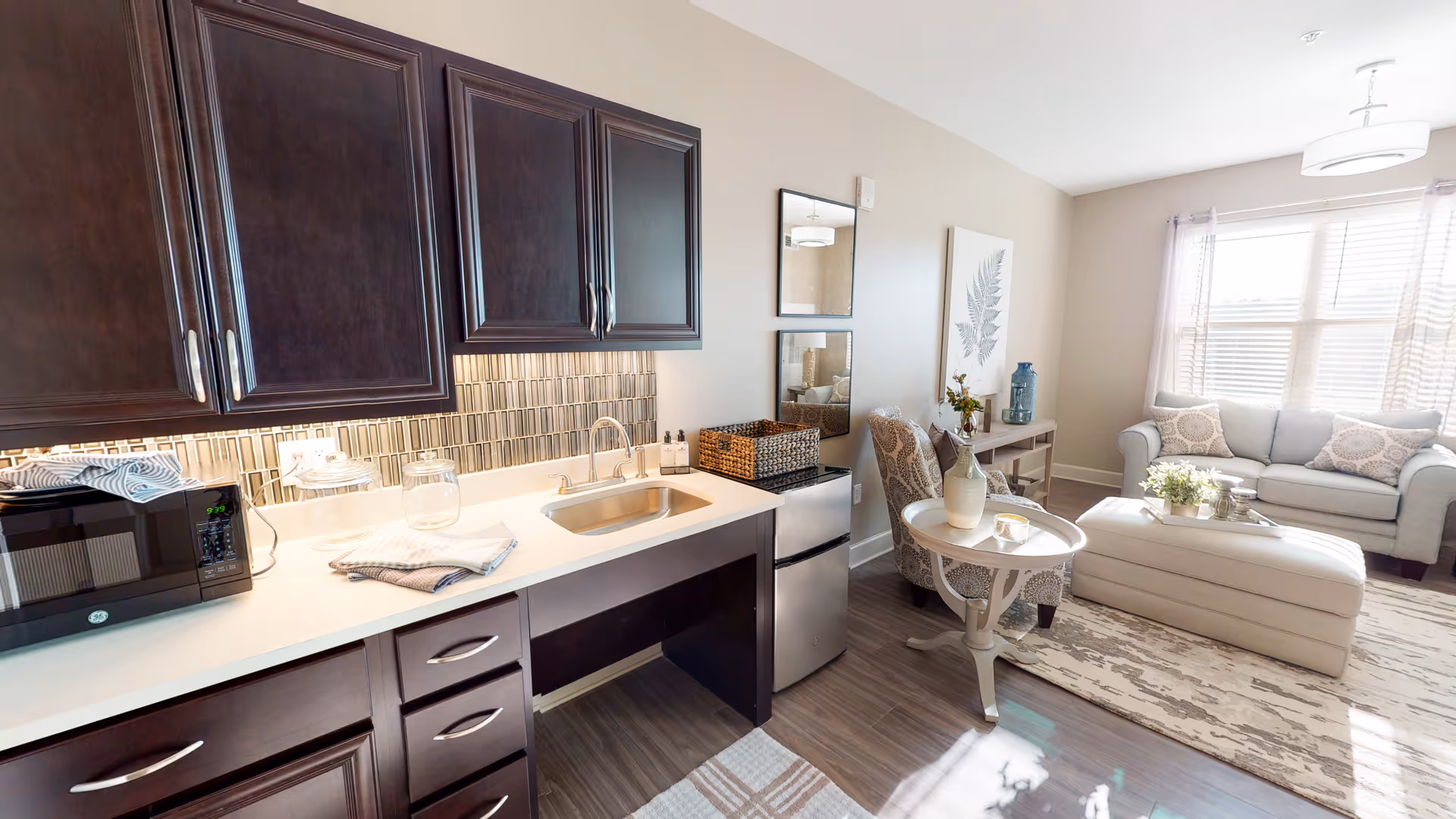 A bright and cozy living space featuring a kitchenette with dark wood cabinets, a small sink, a microwave, and a mini refrigerator. Adjacent to the kitchenette is a sitting area with a patterned armchair, a round side table with a vase, a large ottoman, and a light-colored sofa near a window with sheer curtains. The room has wood flooring and neutral-colored walls with decorative artwork and mirrors.