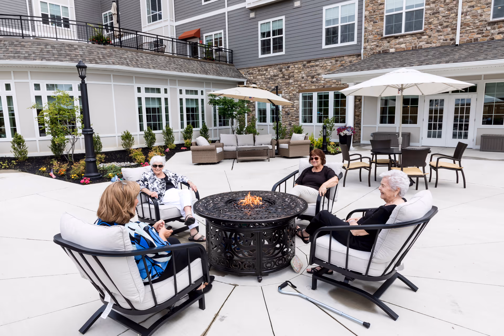 Four elderly women sitting around a decorative outdoor fire pit on cushioned patio chairs in a courtyard of a senior living facility. The courtyard has umbrellas, additional seating, plants, and a building with large windows and stone and siding exterior walls in the background.