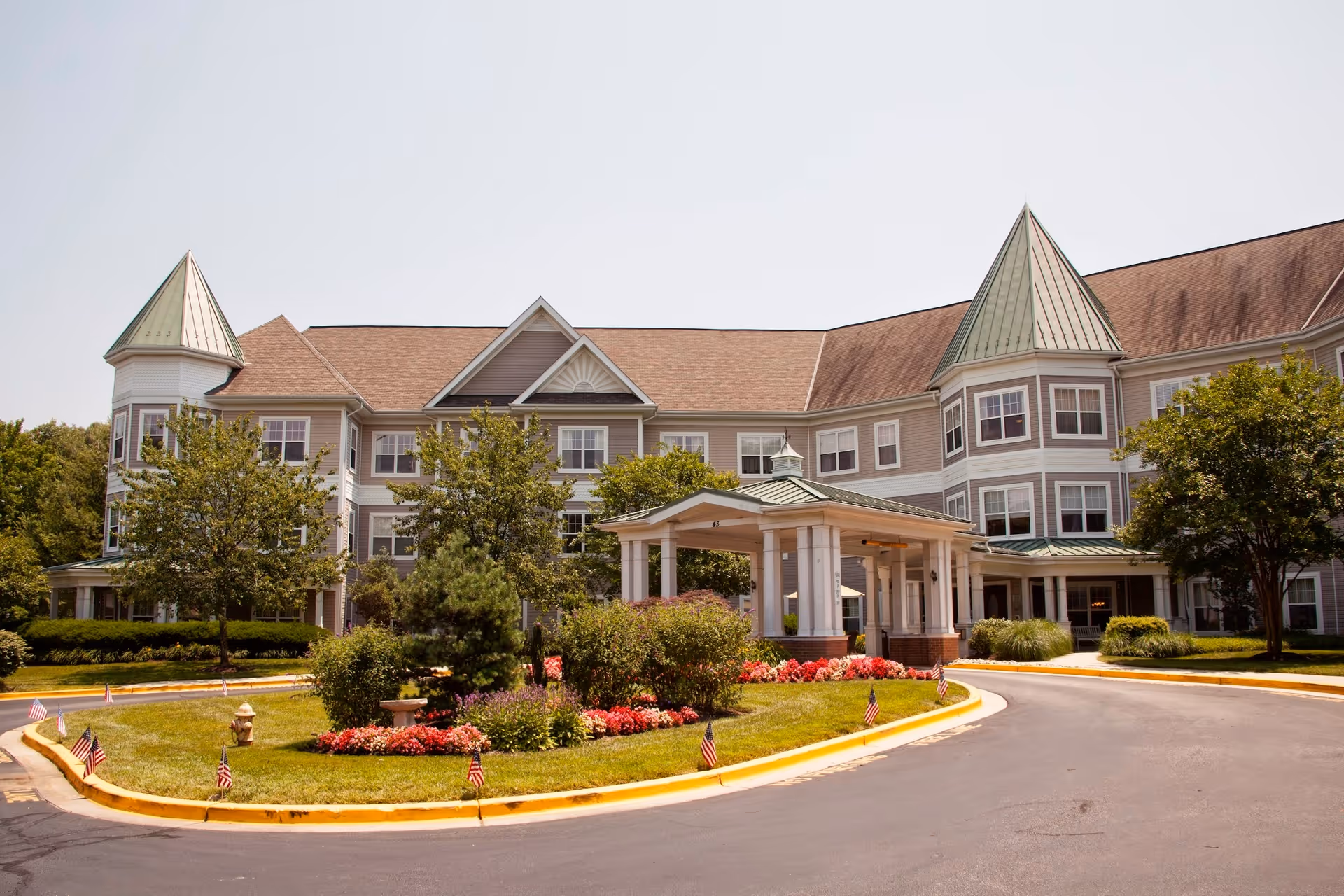 Exterior view of a large senior living facility building with a covered entrance, manicured landscaping including trees, bushes, and flower beds, and a curved driveway with small American flags along the edge.