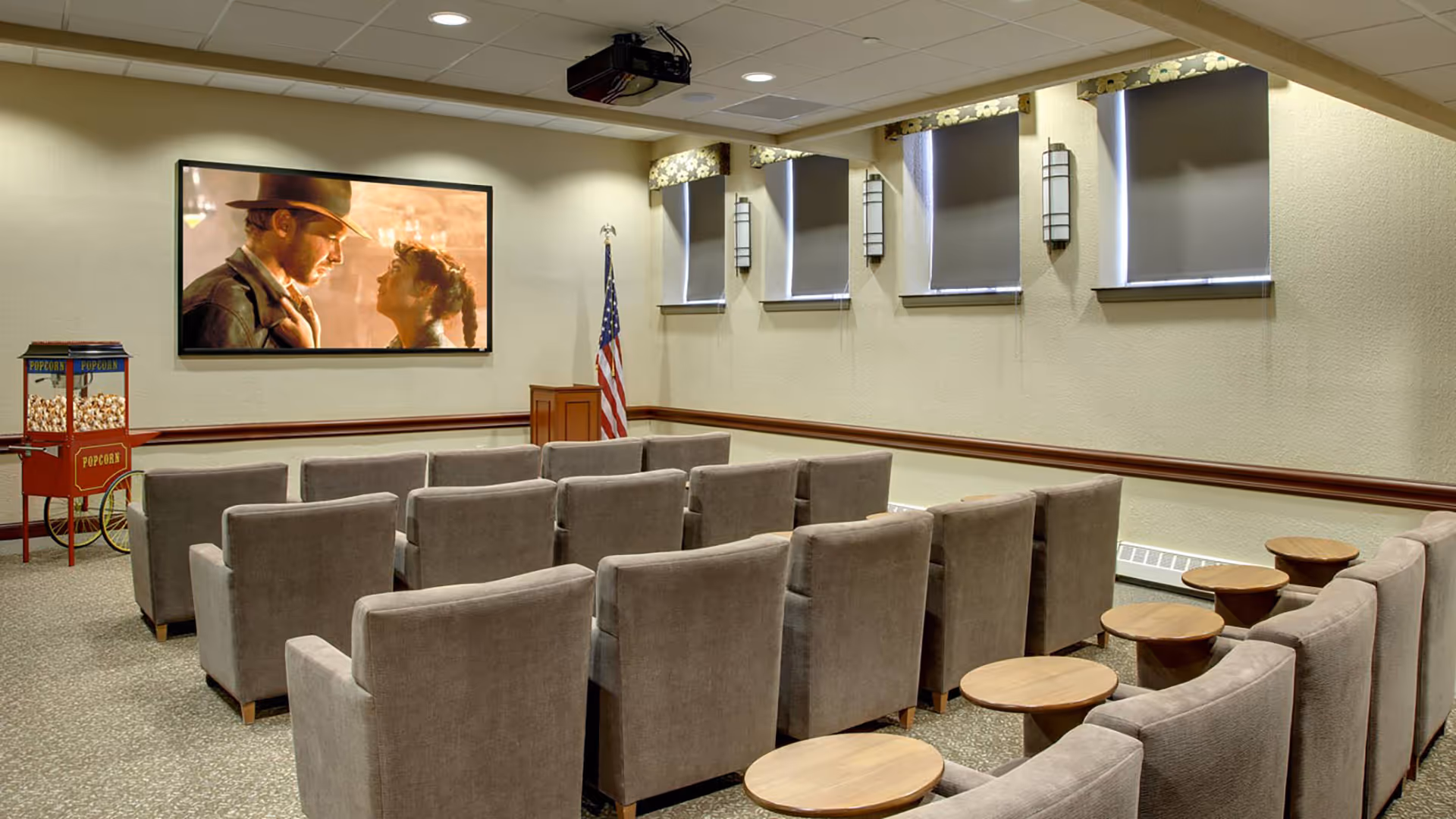 A small theater room with rows of beige upholstered chairs facing a large screen showing a movie scene. There is a popcorn machine on the left side and an American flag near the front. The room has several windows with dark blinds and wall-mounted lights.