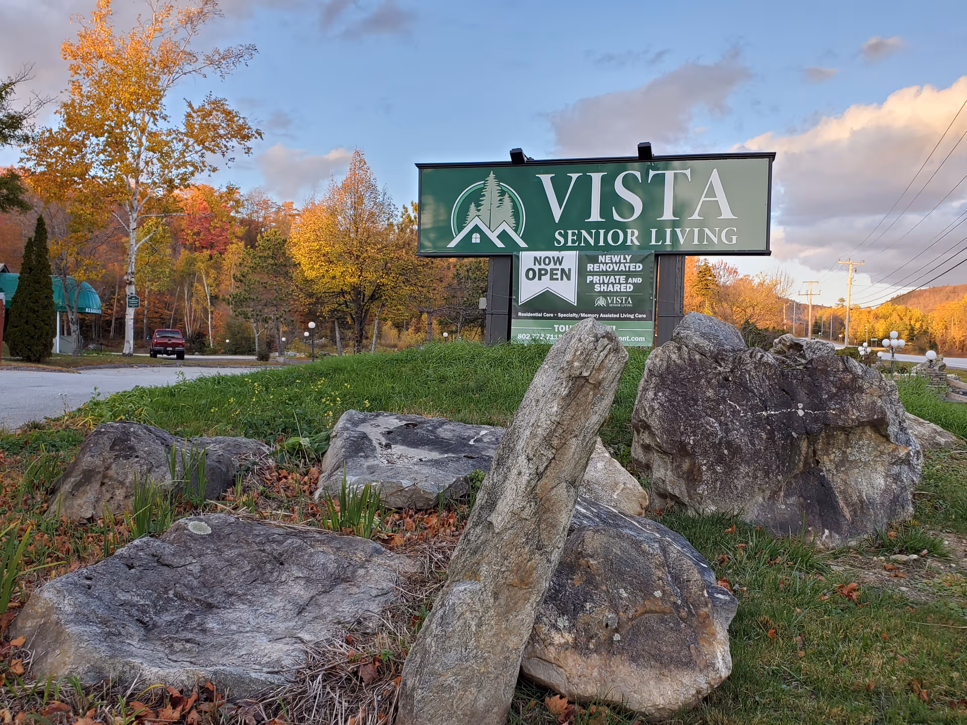 A roadside sign for Vista Senior Living on a grassy mound with large rocks and autumn trees in the background.