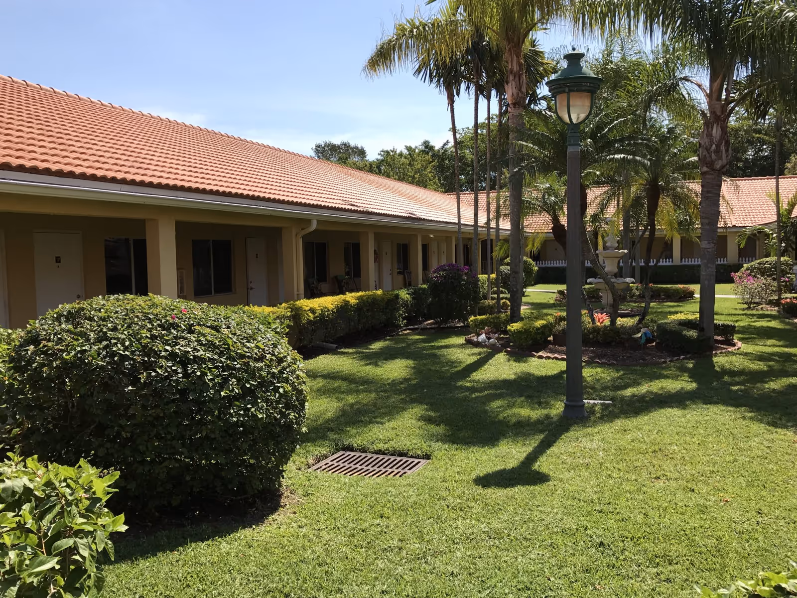 Outdoor courtyard area of a senior living residence with well-maintained green grass, bushes, palm trees, a decorative fountain, and a lamp post. The building has a tiled roof and multiple doors and windows facing the courtyard.