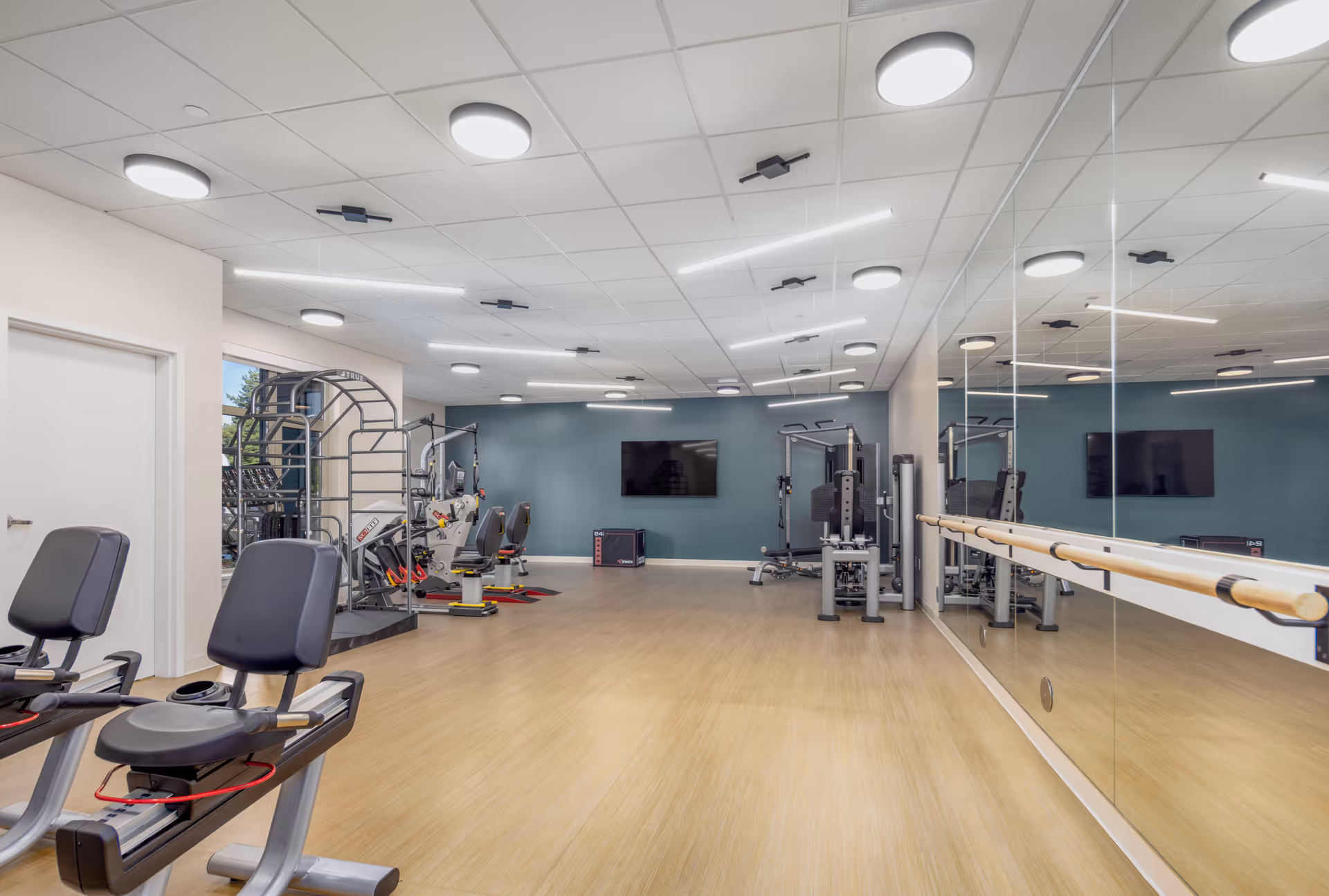 A spacious fitness room with exercise equipment including recumbent bikes, weight machines, and a large wall-mounted TV. The room has a light wood floor, a mirrored wall with a ballet barre, and modern ceiling lights.