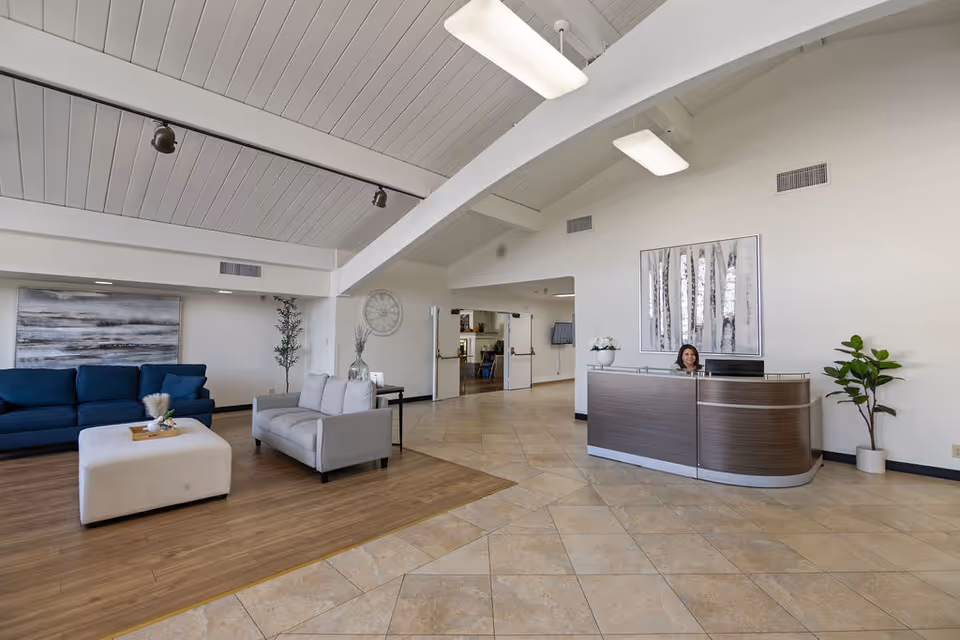 A spacious reception area with a modern wooden reception desk where a woman is seated. The room features a high white ceiling with exposed beams, tiled flooring near the desk, and wooden flooring in the seating area. The seating area includes a blue couch, a gray loveseat, and a white ottoman with a decorative tray. There are plants and wall art decorating the space, creating a welcoming atmosphere.