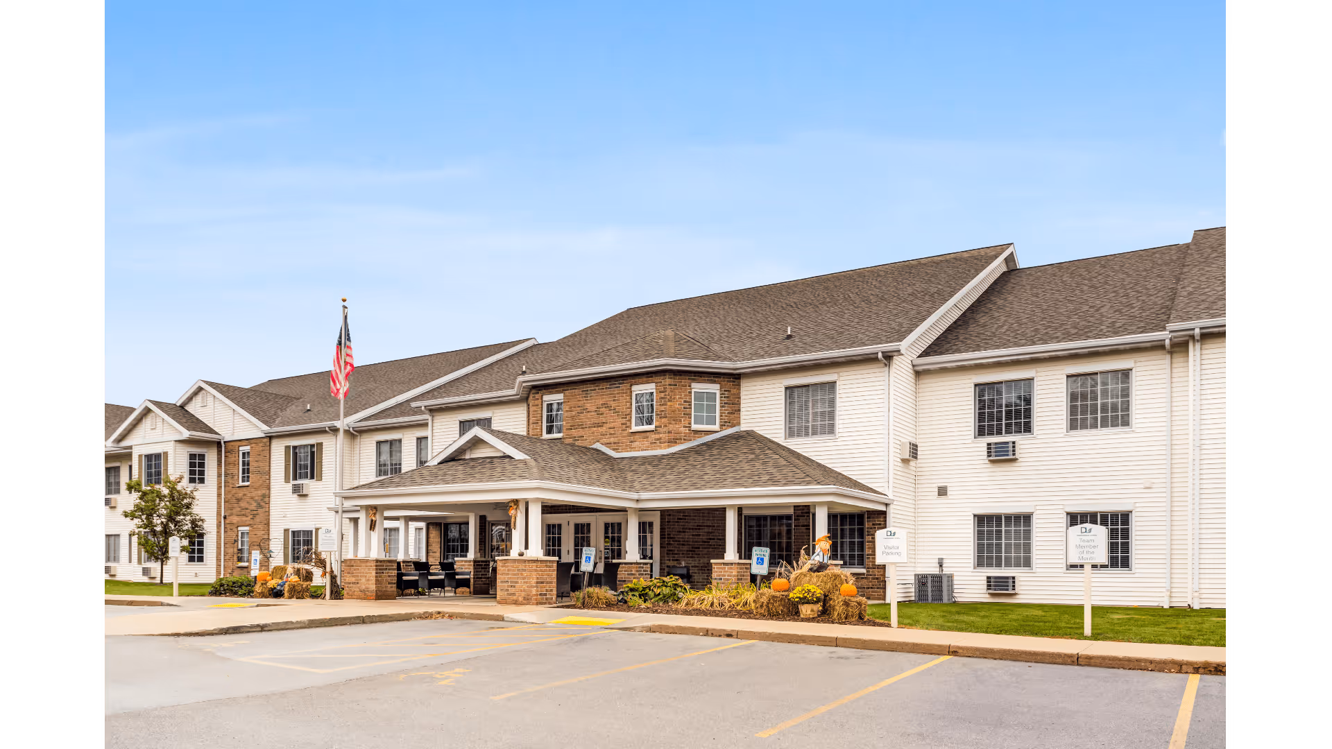 Front exterior of a two-story senior living facility with an entrance canopy, American flag, seasonal pumpkins, and a parking area.