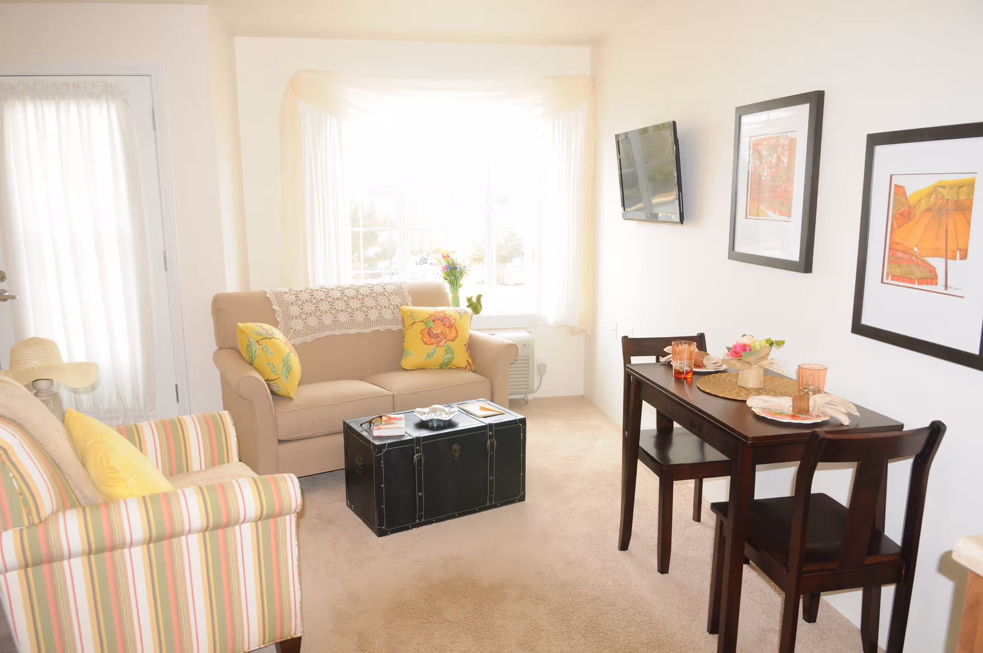 A bright and cozy living room with a beige loveseat adorned with yellow floral pillows and a lace doily, a striped armchair with a yellow pillow, a black trunk used as a coffee table, a small dark wood dining table set for two with pink glasses and a flower centerpiece, a wall-mounted TV, and two framed artworks on the wall. Light streams in through a large window with sheer curtains.