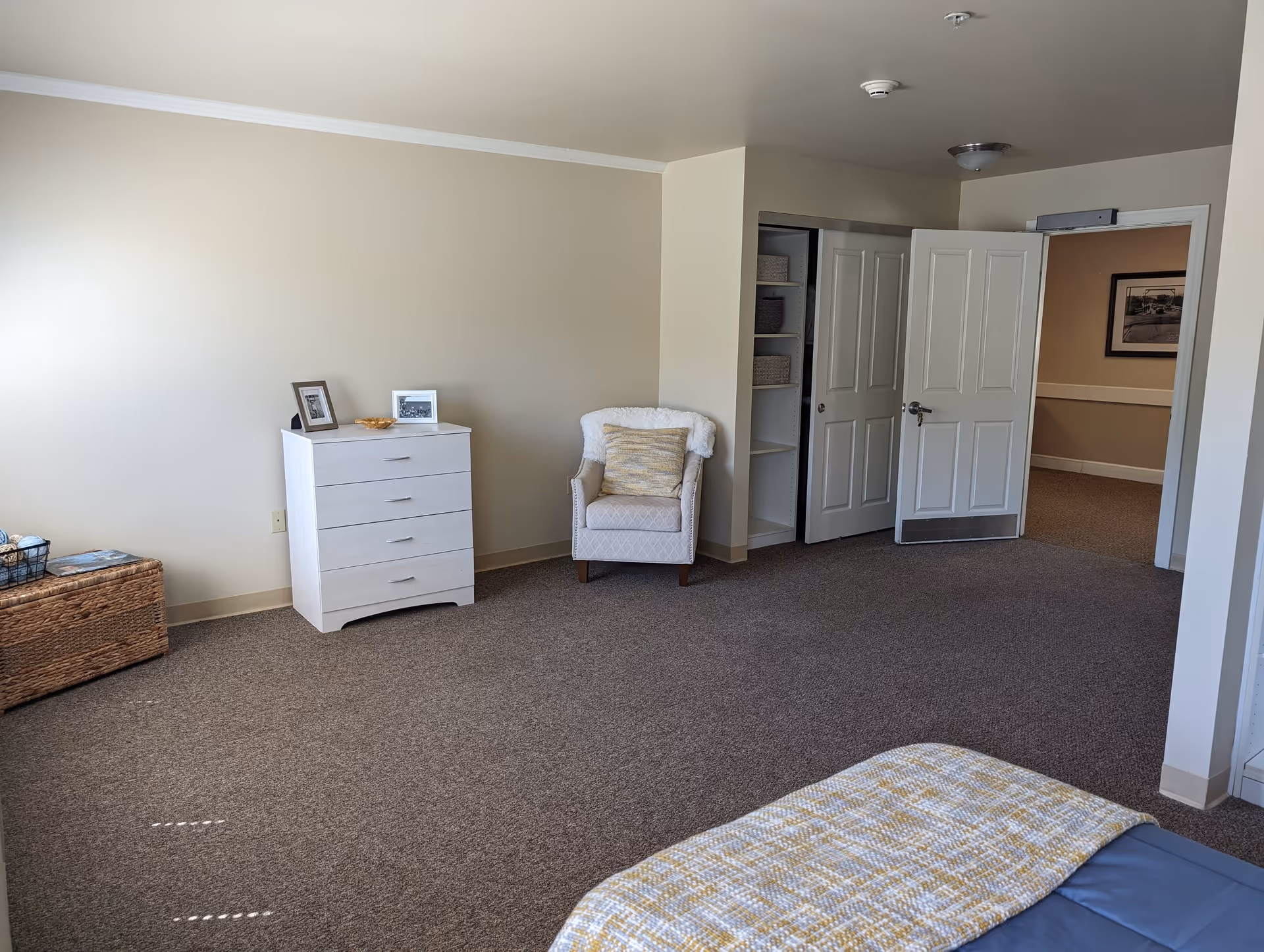 A spacious bedroom in a memory care facility with beige walls and carpeted floor. The room features a white chest of drawers with two framed photos and a small bowl on top, a cushioned armchair with a throw and pillow, an open closet with shelves and baskets, and a partially open door leading to a hallway with a framed picture on the wall. The corner of a bed with a blue and yellow patterned blanket is visible in the foreground.
