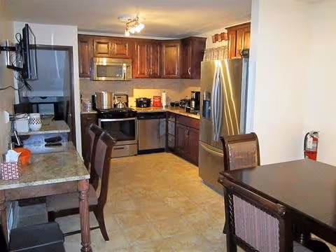A kitchen area with dark wooden cabinets, stainless steel refrigerator, microwave, dishwasher, and stove. There is a dining table with chairs on the right side and a countertop with chairs on the left. The floor is tiled and there is a fire extinguisher mounted on the wall near the dining table.