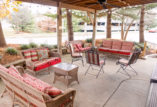 Covered outdoor patio with wicker and metal chairs and sofas with red patterned cushions arranged around a central table under string lights.
