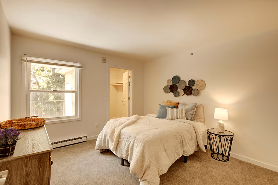A cozy bedroom with a neatly made bed featuring multiple pillows and a light-colored comforter. There is a decorative wall art above the bed and a small round side table with a lamp on the right side. A wooden dresser with a basket and a small potted plant is on the left side near a window with a view of trees outside. An open door reveals a walk-in closet.