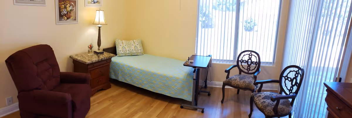 A cozy room with a single bed covered with a blue and yellow patterned bedspread, a wooden nightstand with a lamp and small decorative items, a maroon armchair, two ornate wooden chairs with patterned cushions, and large windows with vertical blinds letting in natural light.