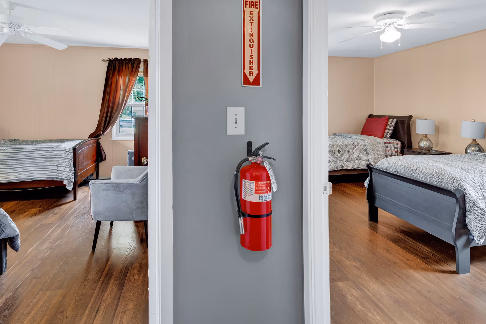 View of two adjacent bedrooms separated by a wall with a mounted red fire extinguisher and a fire extinguisher sign above it. Each bedroom has a bed with patterned bedding, a ceiling fan, and wooden flooring. One bedroom has a window with brown curtains and a gray armchair.