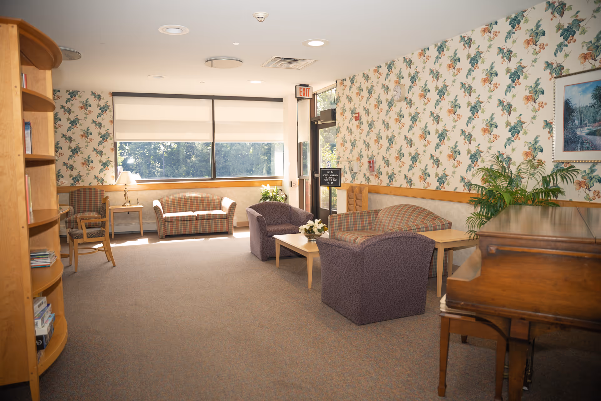 Sunlit common sitting area with armchairs, sofas, a coffee table, bookshelf and piano against floral wallpaper.