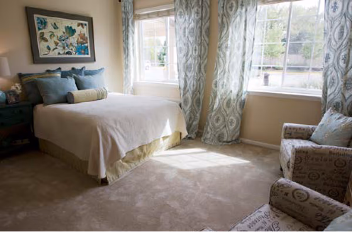 A bright bedroom with a neatly made bed featuring multiple pillows, a framed floral artwork above the bed, two large windows with patterned curtains letting in natural light, and two upholstered armchairs with decorative pillows.