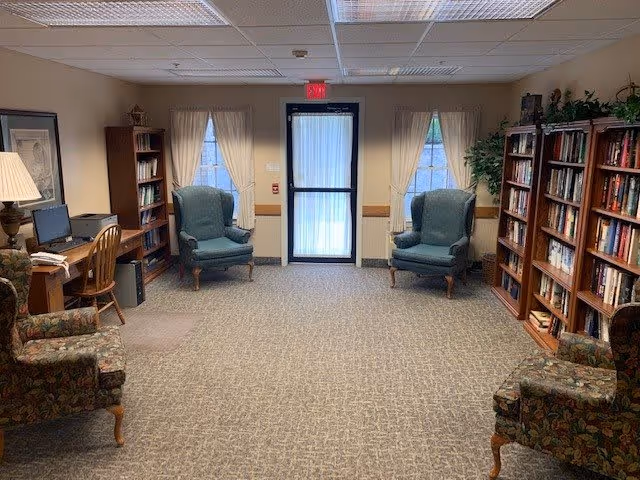 A cozy interior room with two blue armchairs placed near a door with windows on either side covered by light curtains. The room has bookshelves filled with books on both sides, a wooden desk with a computer and chair on the left, and two floral patterned armchairs in the foreground. The ceiling has fluorescent lights and there is a carpeted floor.