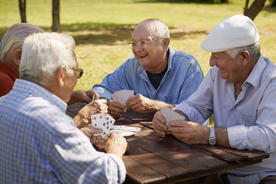 Four elderly men sitting around a wooden picnic table outdoors, playing cards and smiling, with grass and trees in the background.