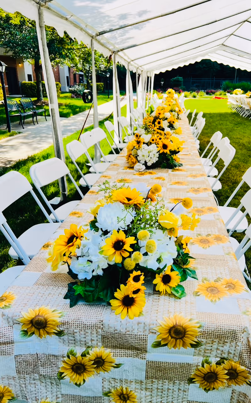 Long outdoor table covered with a sunflower-themed tablecloth and decorated with multiple sunflower and white flower centerpieces under a white canopy tent, surrounded by white folding chairs on a grassy lawn near a building.